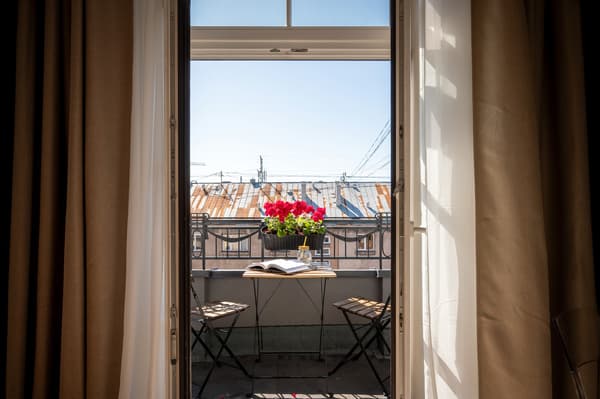 Small balcony with two chairs, a table with flowers and an open book, seen through open curtains and a glass door.