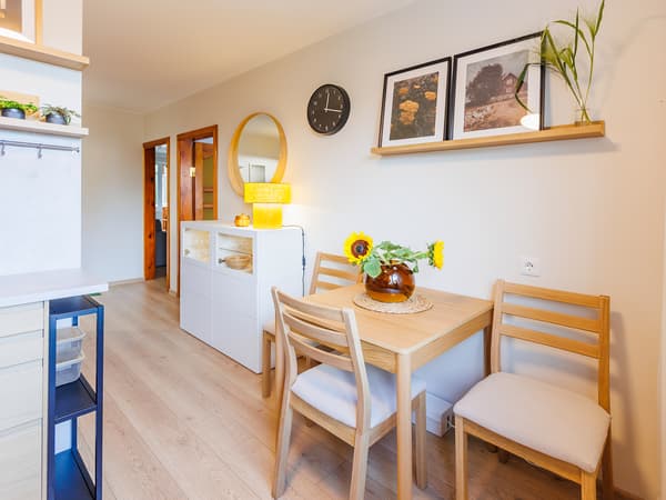 Small dining area in a modern kitchen with a wooden table, two chairs, a vase of sunflowers and framed pictures on a wall shelf.