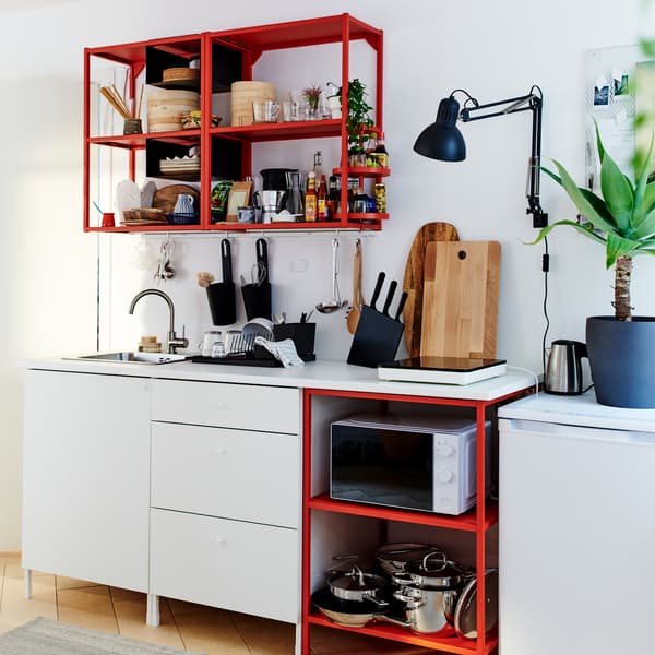 Small kitchen in red/white with a portable induction hob, wooden chopping boards, a black wall lamp and a black dish drainer.
