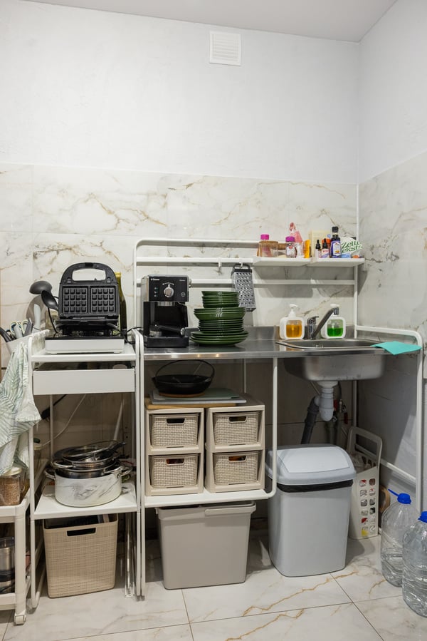 Small kitchen with open metal shelving, plastic storage boxes and appliances on the worktop; a corner sink and various storage solutions are visible.