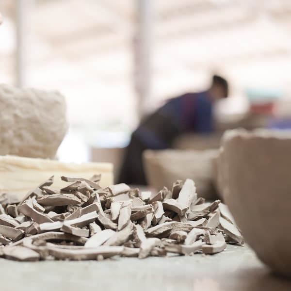 Small pieces of broken pottery on a table with larger intact ceramics and an artisan in the background.