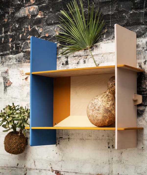 Small, rectangular wall shelf made of plywood, partly painted in yellow and blue, mounted on a rough brick wall.