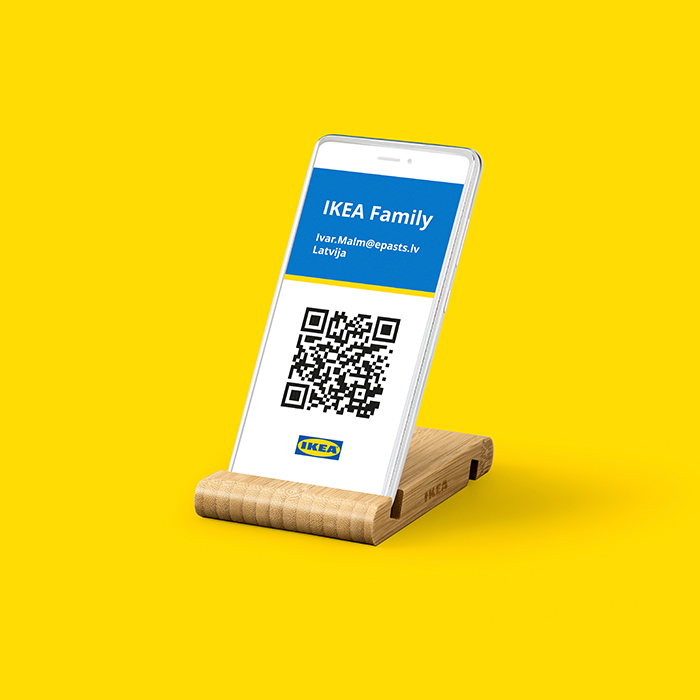Smartphone displayed on a wooden IKEA stand, showing an IKEA Family digital membership card with a QR code, Lithuanian email address, and the word 'Lietuva', set against a bright yellow background.
