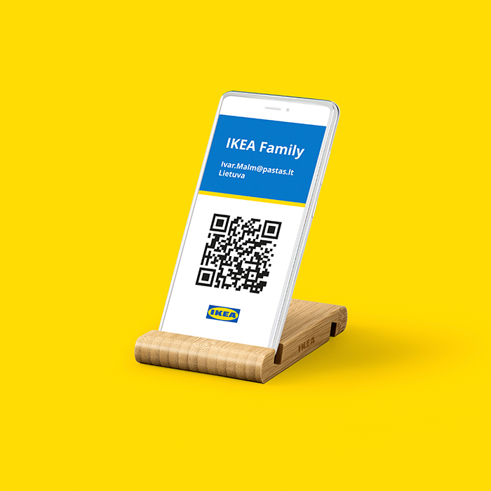 Smartphone displayed on a wooden IKEA stand, showing an IKEA Family digital membership card with a QR code, Lithuanian email address, and the word 'Lietuva', set against a bright yellow background.