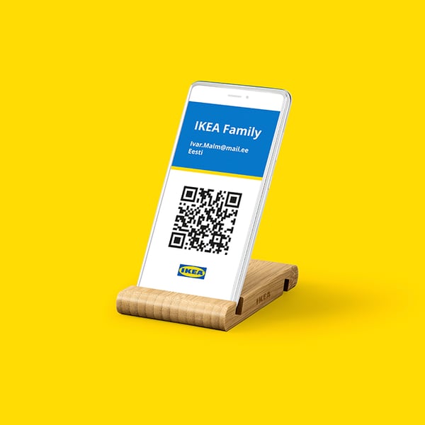 Smartphone displaying an IKEA Family digital membership card with a QR code, placed on a wooden stand against a bright yellow background.