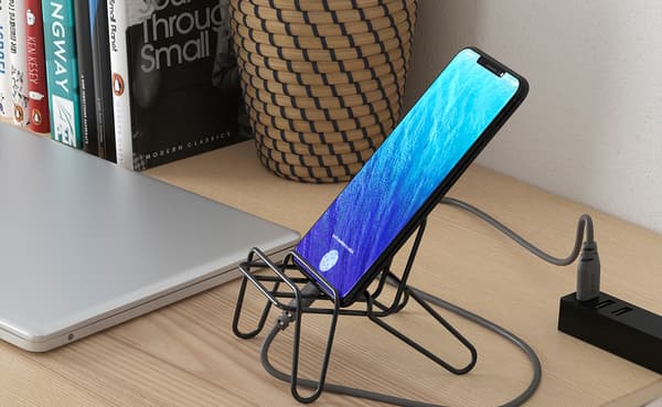 Smartphone with a vivid blue screen rests on a black stand, charging beside a laptop and power strip on a warm wooden desk.