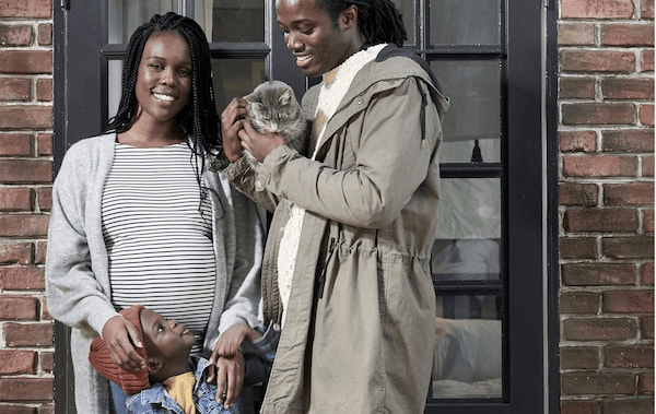Smiling, late-twenties man and pregnant woman in front of a brick wall, a small boy in the middle; the man holds a cat.