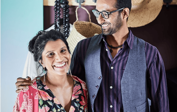 Smiling, thirty-something woman and man standing affectionately together; clothing and accessories on a rack behind them.