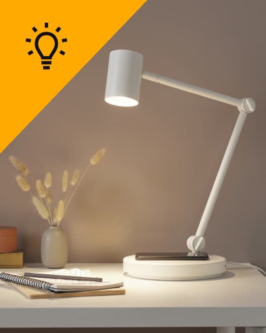 Sobre un escritorio blanco de IKEA una lámpara de escritorio de cabezal retractil encendida.