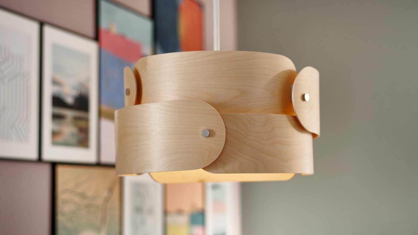 מנורת תלייה SÖDÅKRA, העשויה מעץ תרזה, עם שכבות עץ המונחות זו מעל זו, על רקע של קיר תמונות ססגוני.