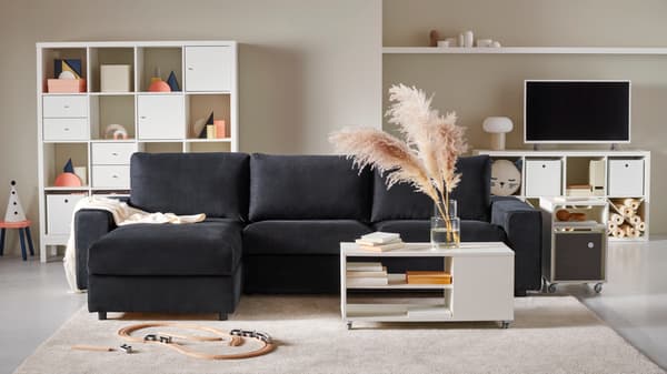 Sofá azul marino VIMLE en un living con tonos claros o pasteles, como el blanco y rosado pálido. Atrás de el hay muebles blancos llenos de decoración, libros y un televisor con una lámpara gris con blanco.
