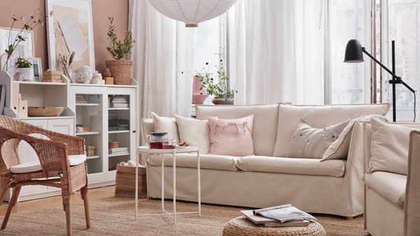 Sofá beige BACKSÄLEN con cojines rosados y cafe claro. Todo en un living con colores tierra y una silla con fibras naturales y un mueble blanco donde se guardan libros y decoración.