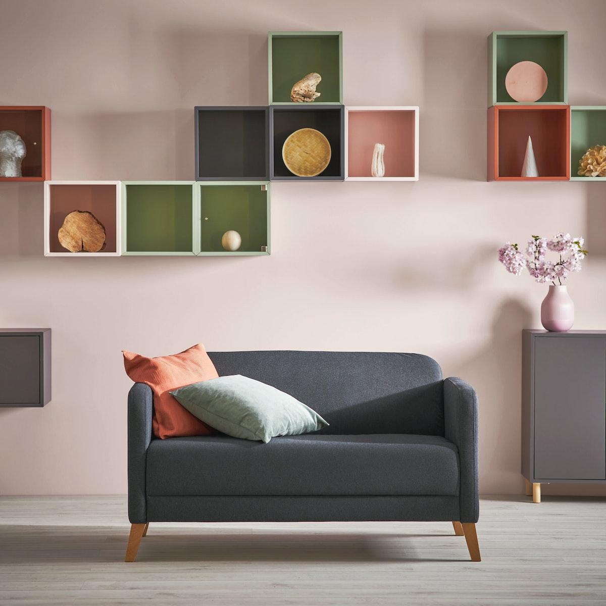 Sofá de la serie LINANÄS de color gris oscuro co ndos cojines encima, detras de él una pared de color rosado pálido con una serie de estantes modulares que contienen accesorios decorativos.