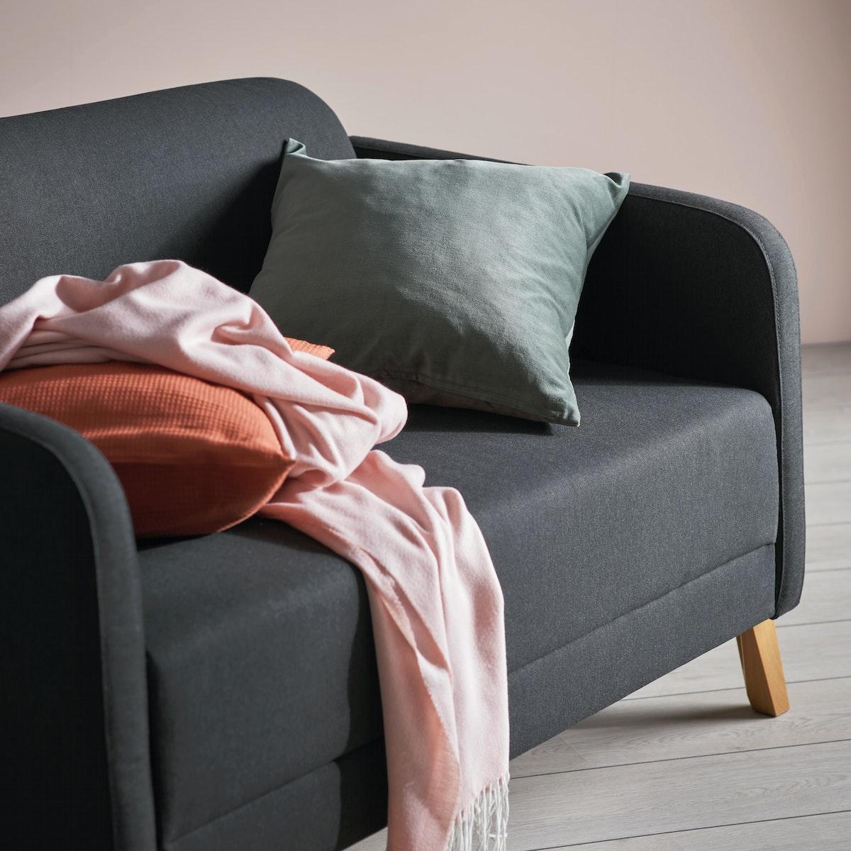 Sofá de la serie LINANÄS de color gris oscuro,tras de el una pared rosada pálido. Mantas y cojines sobre el sofá.