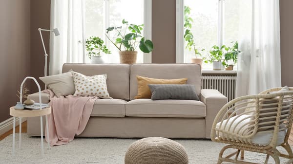 Sofa KIVIK z beżowym pokryciem staojąca w jasnym pokoju dziennym z brązowymi ścianami.