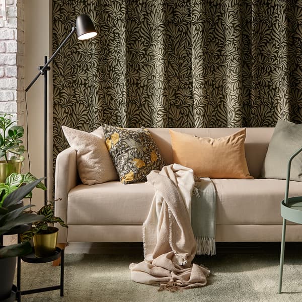 Sofa LINANÄS berwarna kuning air dengan pelbagai kusyen terletak di atas ambal berwarna kelabu-hijau muda, di hadapan langsir bercorak daun yang tergantung pada dinding.