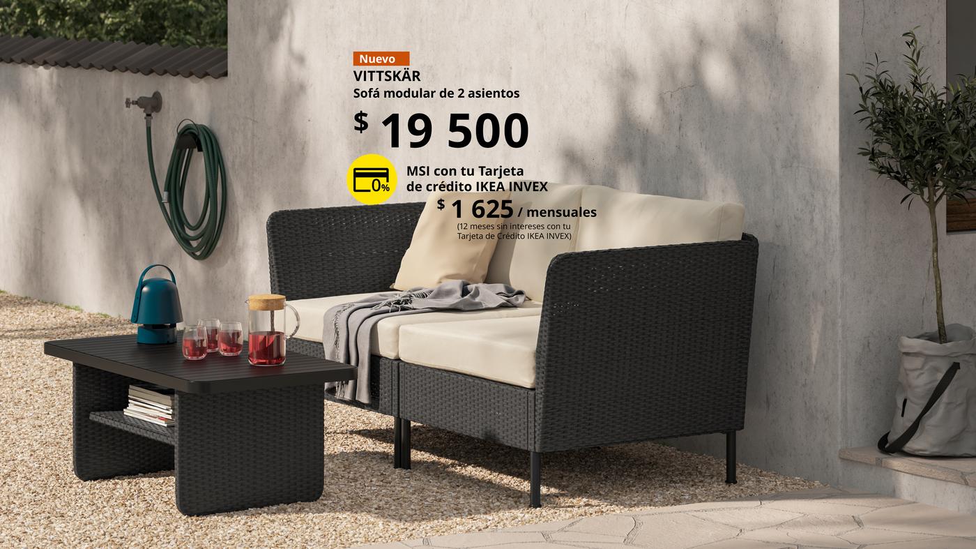 Sofá modular VITTSKÄR de 2 asientos, elaborado de mimbre de plástico en color oscuro con cojines claros, colocado en un espacio exterior con una mesa auxiliar y elementos decorativos.