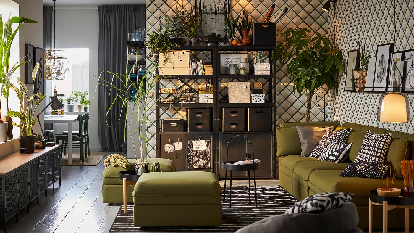 Soggiorno con sistema componibile BROR nero di IKEA, con scatole e contenitori sui 3 ripiani e mobili con ante neri, un divano, due pouf, un tappetp scuro, un tavolino e Mobile TV FJÄLLBO con piante sopra.
