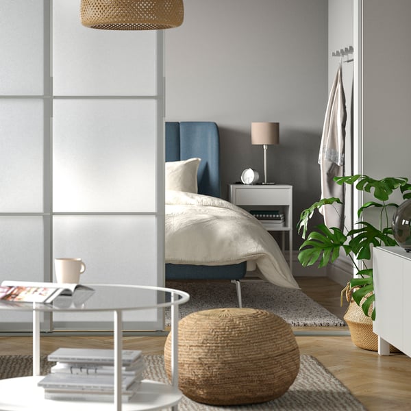 Soggiorno con vista camera da letto. Nel soggiorno ci sono un pouf SANDARED e un tavolino VITTSJÖ bianco. Nella camera da letto ci sono una struttura letto imbottita e un comodino con sopra un lampada da tavolo.