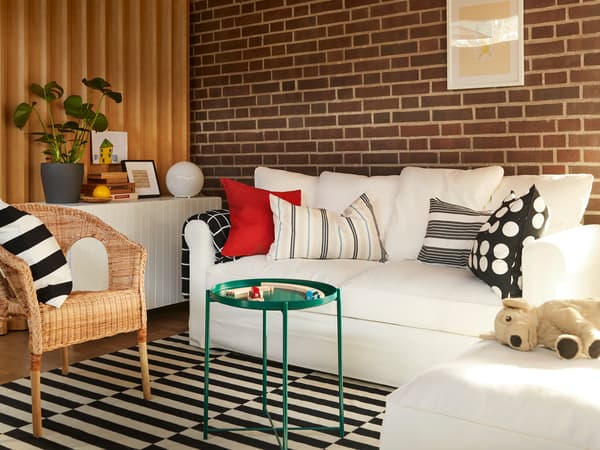 Soggiorno moderno con divano letto GRÖNLID color crema con chaise-longue su un tappeto STOCKHOLM a righe. Al centro della stanza un tavolino verde con una poltrona con cuscino AGEN. Sull'angolo della stanza un mobile bianco con sopra un vaso, una foto, una lampada e un dipinto.