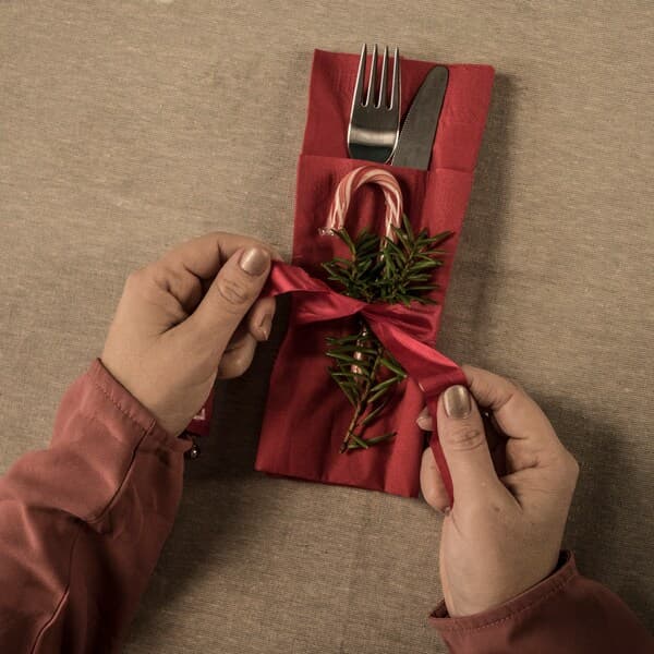 Someone decorates a napkin bag containing a knife and fork with a bow, a candy cane and a fir branch