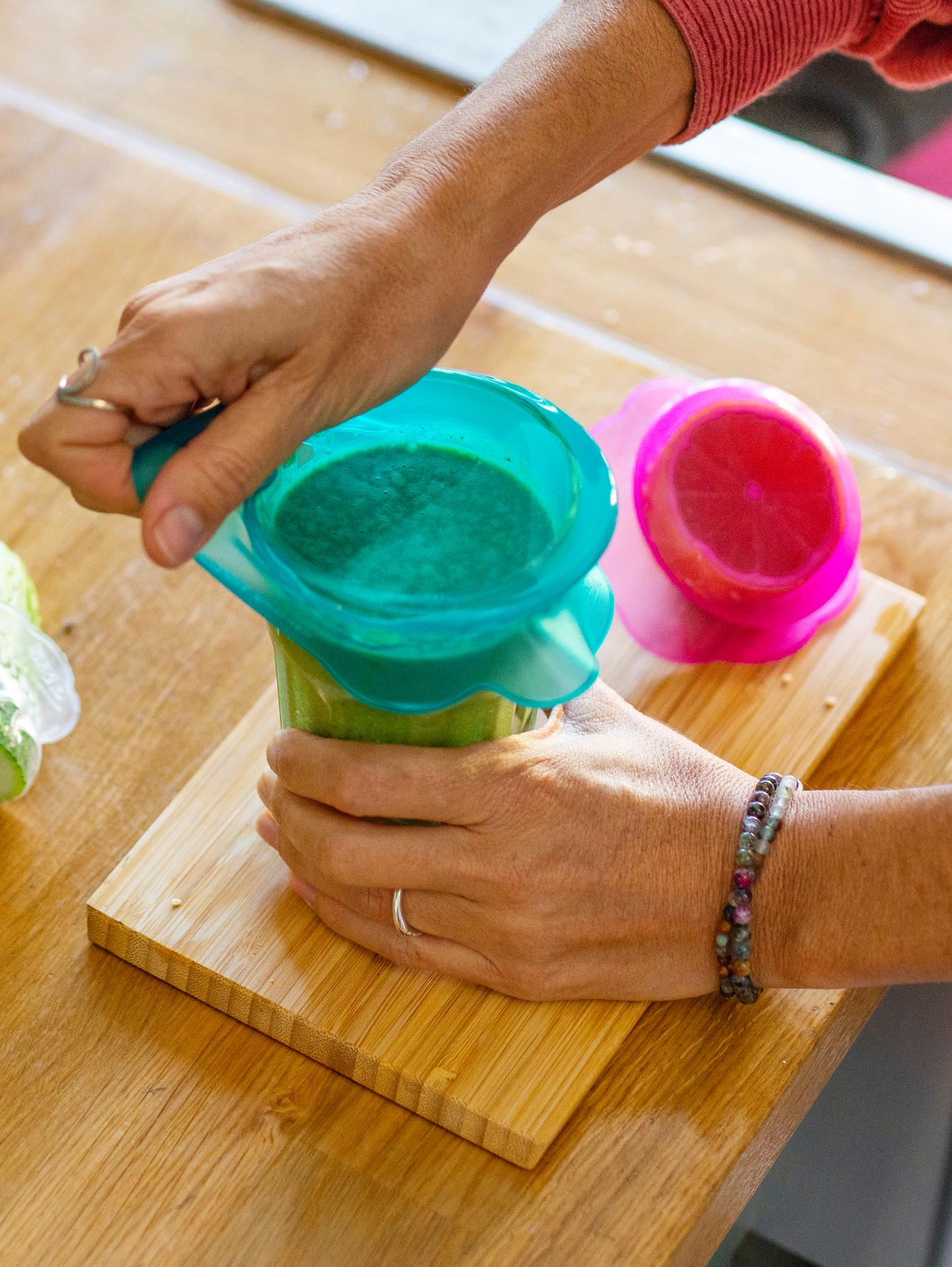 Someone using a turquoise, silicone ÖVERMÄTT food lid on a glass with watermelon pieces placed on a cutting board.