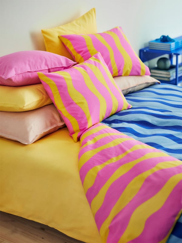 SOMMARVICKER duvet and two pillows in pink and yellow stripes are placed on a bed covered with a yellow NATTSVÄRMARE bed cover. Four other pillows in yellow and pink NATTSVÄRMARE covers are propped against the wall. 