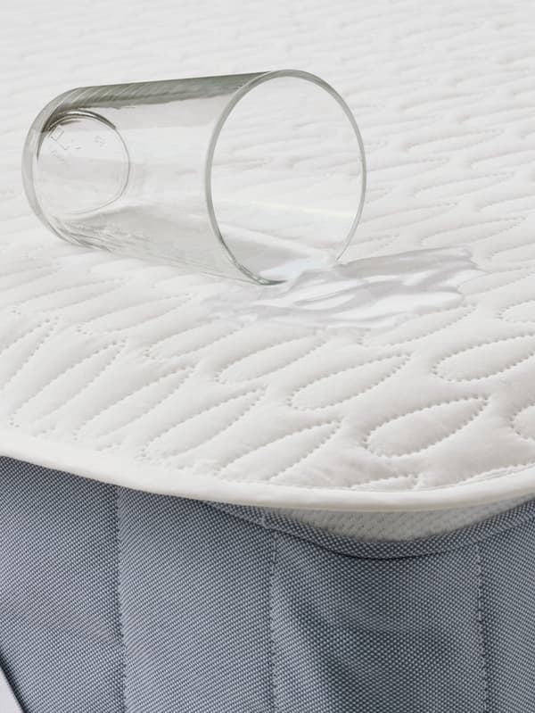 SOTNÄTFJÄRIL mattress protector is laid on a mattress with a glass of water being spilled on the mattress, showing its waterproof protection. 