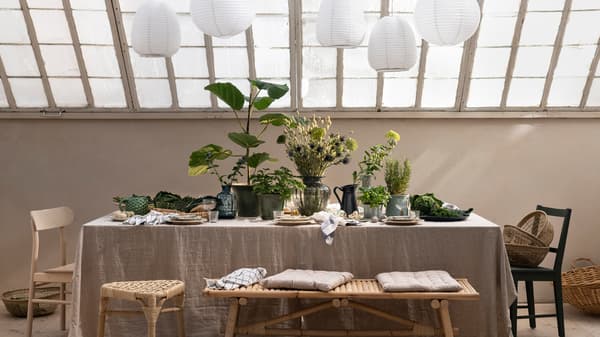 Sous un toit de verre, des assiettes, des plantes et des feuillages dans des cache-pots, ainsi que des vases sont disposés sur une table munie d’une nappe en lin.
