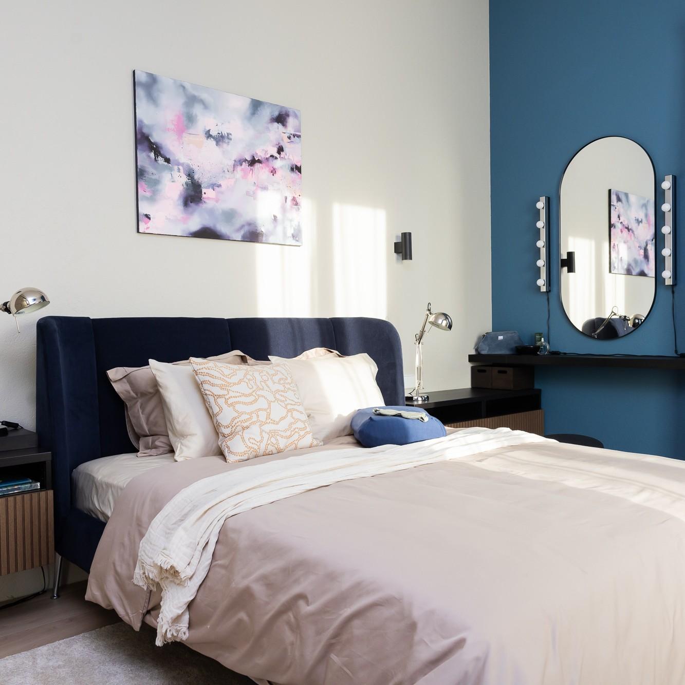 Современная спальня с тёмно-синим мягким изголовьем, светлым постельным бельём и декоративными подушками; над кроватью — абстрактная картина, в углу — синяя акцентная стена с круглым зеркалом и небольшим столиком.
