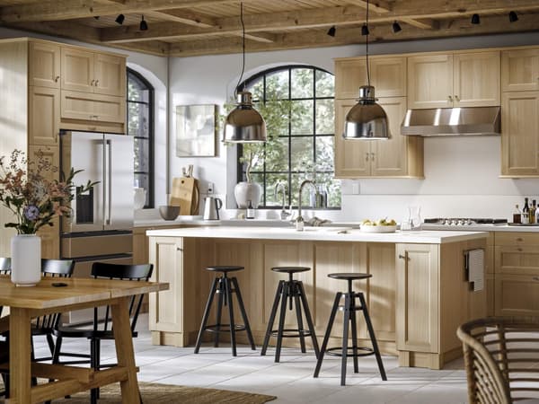Spacious and well-lit modern kitchen with wooden cabinets, a central island with bar stools, stainless steel appliances, decorative pendant lighting, and a small wooden table in the background decorated with a vase of flowers.