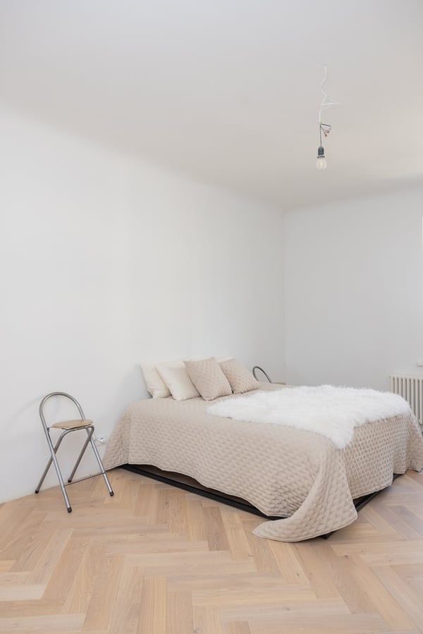 Spacious bedroom with a bed, light-coloured walls and a wooden parquet floor.