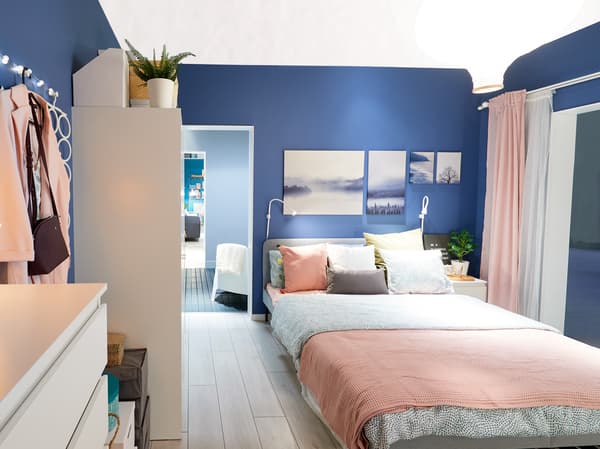 Spálňa s modrou stenou s obrazmi, posteľ s ružovým povlečením a spálňový nábytok v bielej farbe.