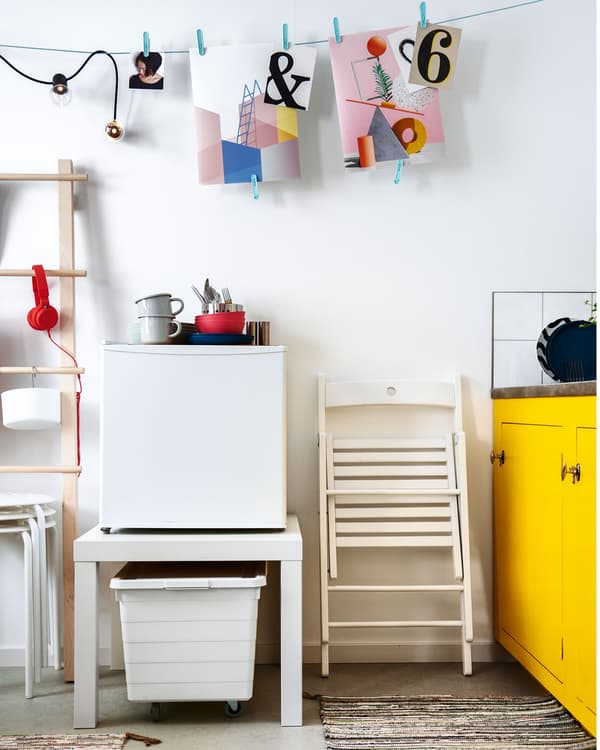 Spațiul de lângă chicinetă cu decorațiuni de perete suspendate de o sfoară. Un frigider mic așezat pe o masă LACK, un coș dedesubt.