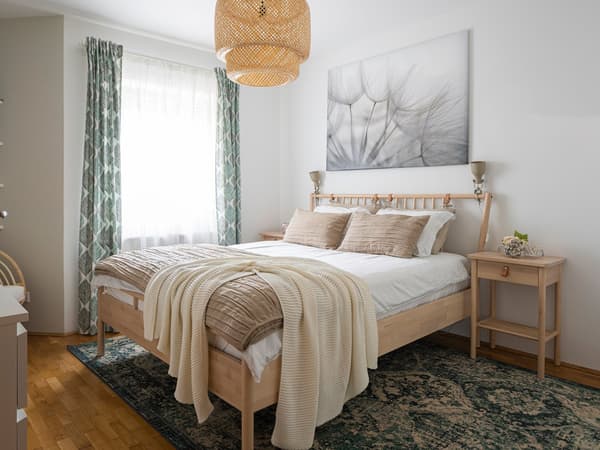 Spavaća soba s drvenim krevetom, bijelom posteljinom, smeđim jastucima, pletenom lampom i slikom maslačka na zidu.