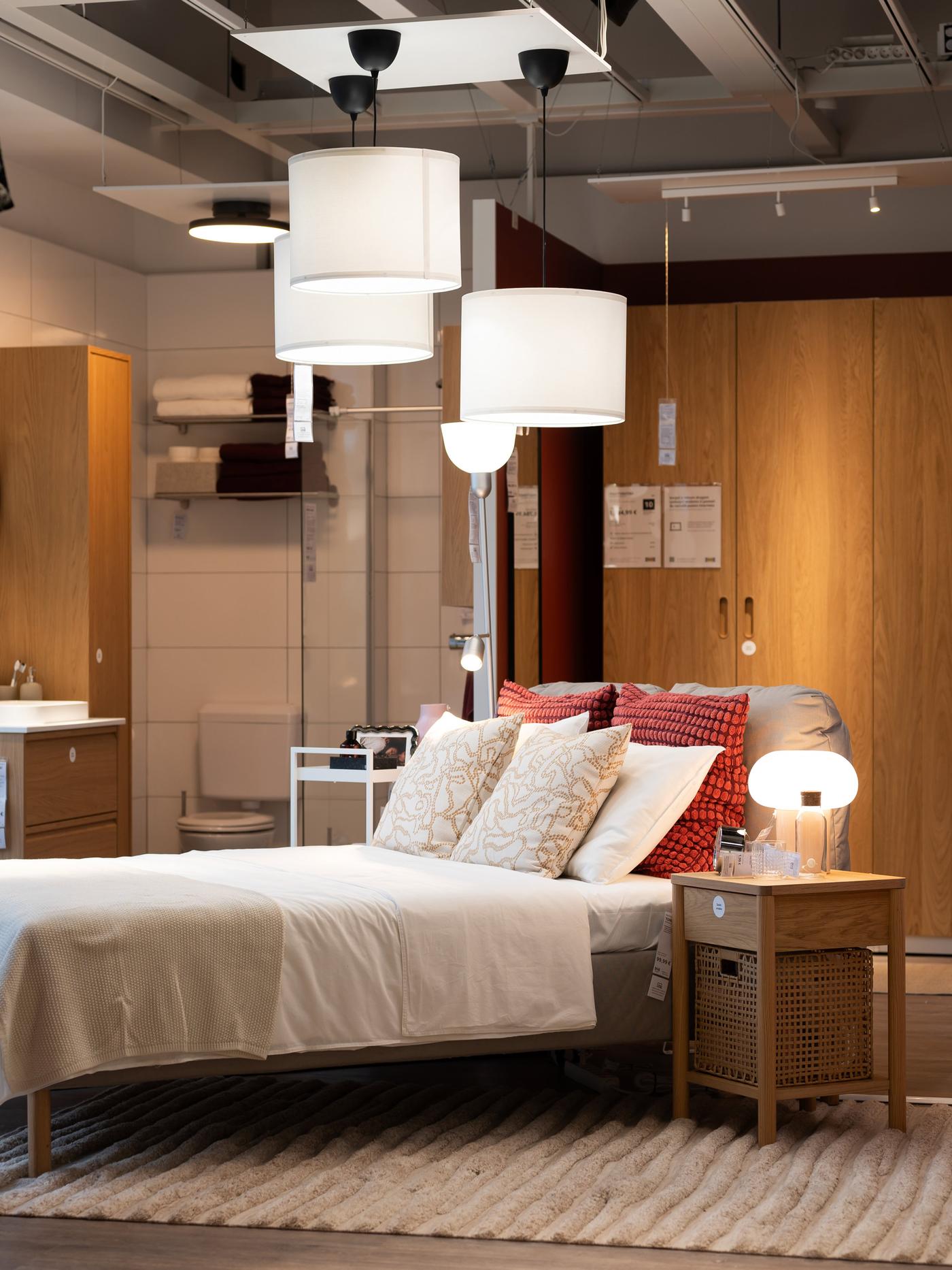 Spavaća soba s krevetom, noćnim ormarićem, lampom i ormarima u pozadini. Krevet ima bijelu posteljinu i crvene jastuke.