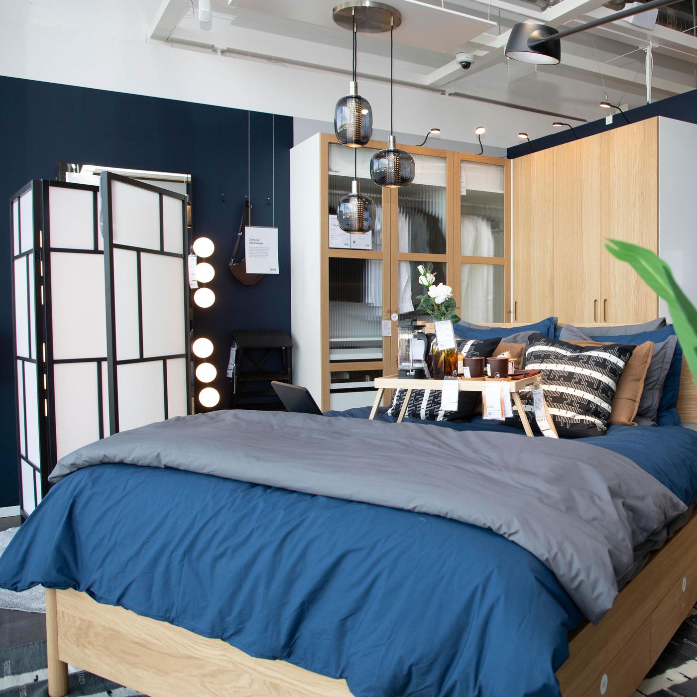 Spavaća soba s plavim krevetom, drvenim noćnim ormarićem, crno-bijelim paravanom i ormarom sa staklenim vratima; na krevetu poslužavnik s cvijećem i pićem, iznad kreveta tri crne viseće lampe.