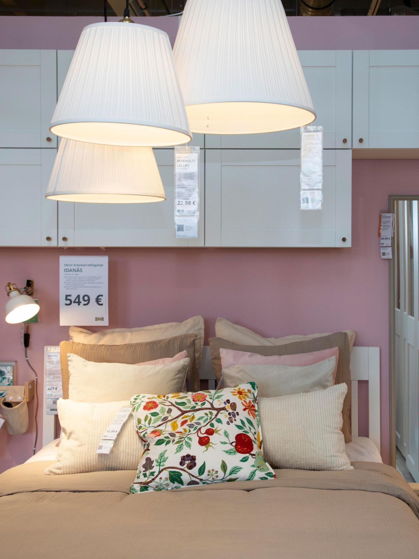 Spavaća soba s ružičastim zidom, bijelim ormarićima, visećim bijelim lampama i krevetom s bež posteljinom i šarenim jastukom; prikazana cijena kreveta je 549 €.