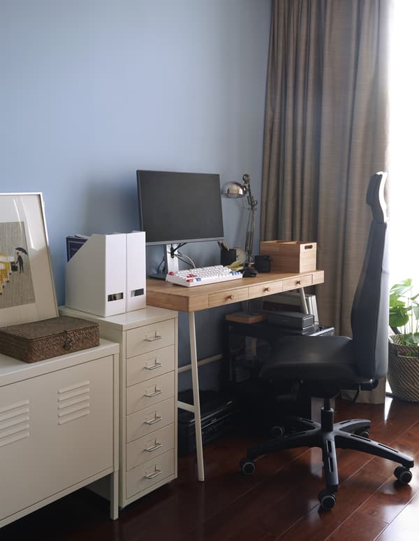 Spazio di lavoro con monitor, scrivania, sedia da gaming, cassettiera HEMNES, mobile bianco sullo sfondo di una parete azzurra. Sotto la scrivania, una stampante.