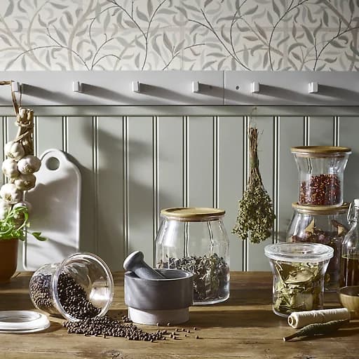 Spices in glass containers with wooden lids on a countertop and herbs hanging from hooks.