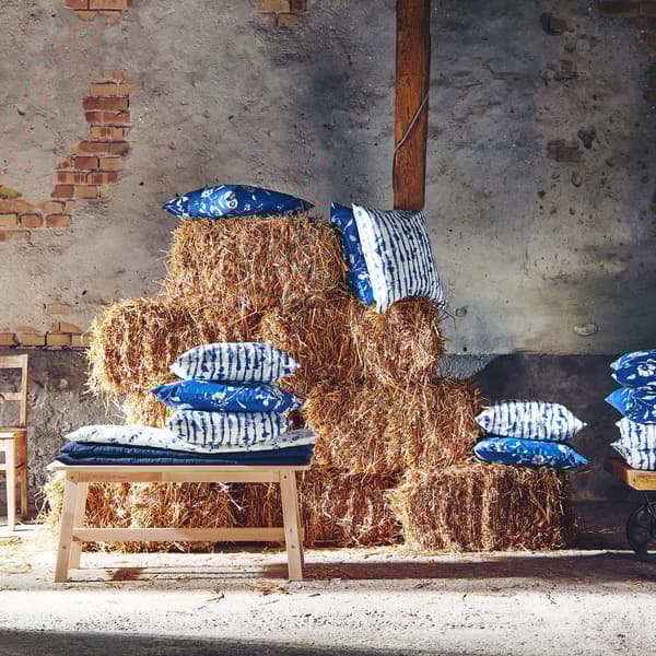 Square, blue-and-white BLÅGRAN and SIMSPORRE cushion covers with floral and dyed patterns are shown on piles of hay.
