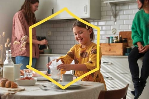 Srečna deklica v rumeni obleki naliva vodo v kuhinji, ob zajtrku. Dve odrasli osebi sta v ozadju. Motiv uokvirja rumen obris hiše.