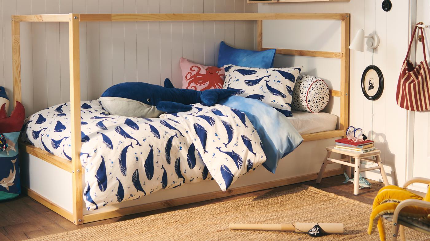 سرير KURA مُزيّن بطقم لحاف حوت BLÅVINGAD أزرق؛ وهناك فوقه دمية ناعمة شكل حوت BLÅVINGAD أزرق.