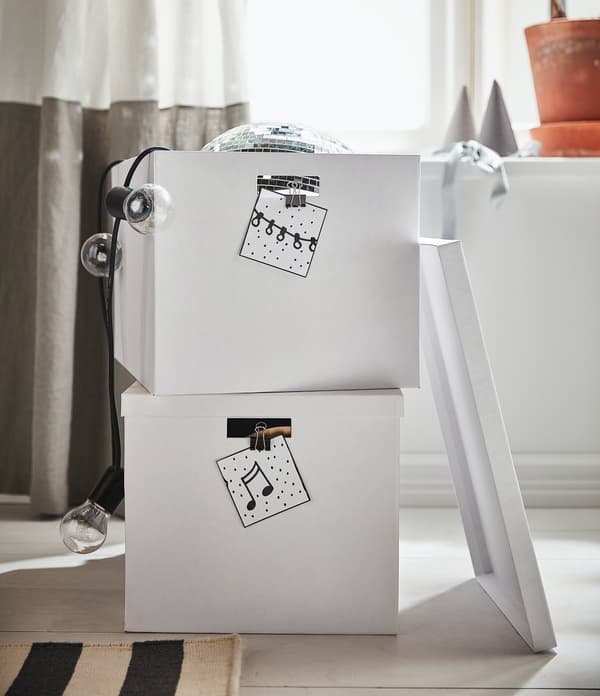 Stacked white boxes with black and white note cards attached to them; a disco ball can be partially seen at the top. Next to them are fairy lights and a potted plant. Cosy and tidy atmosphere.