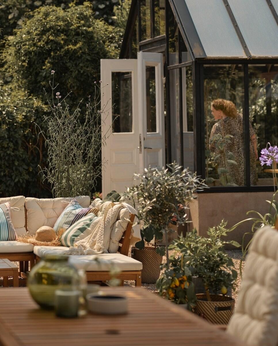 Staklenik s otvorenim vratima, žena unutra, vani vrtni namještaj s jastucima i dekama, biljke u teglama.