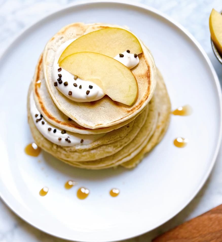 Stapel aus fluffigen Pfannkuchen auf einem weißen Teller, garniert mit Apfelscheiben, Creme und Schokoladenstückchen, mit etwas Sirup darüber.