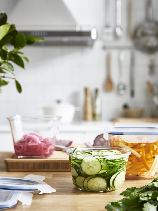 Стеклянные контейнеры для хранения продуктов IKEA 365+, наполненные нарезанными огурцами, луком и другими свежими ингредиентами, расставлены на деревянной кухонной столешнице.