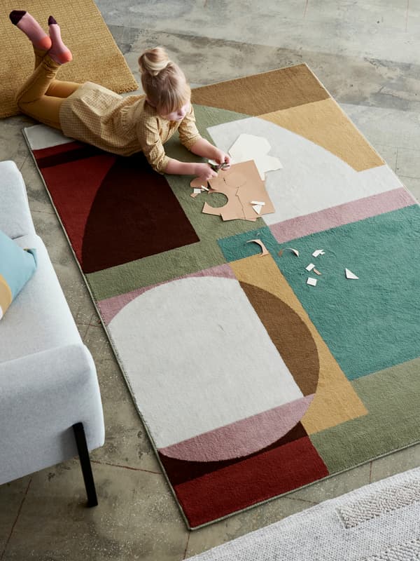 STENMÄTARE rug with a Bauhaus pattern in pastel tones lying by a sofa with a girl on the rug doing arts and crafts.