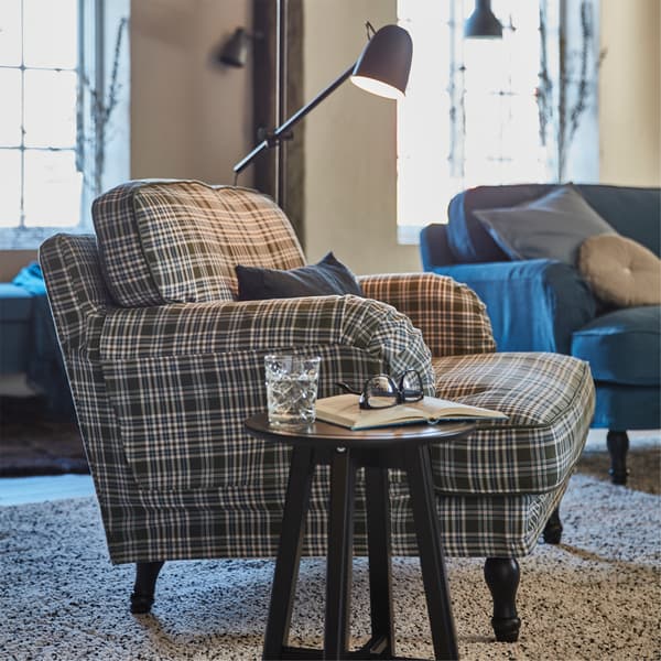STOCKSUND armchair in SEGERSTA multicolour plaid cover with a small table holding a glass, book and eyeglasses, with a floor lamp in the background.