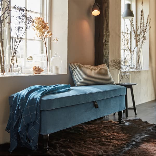 STOCKSUND bench in LJUNGEN blue cover with a pillow, in front of a window with light streaming in and plants on the sill.